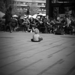 enfant assis de dos en tailleur sur une place, qui regarde des adultes assis en terrasse d'un café